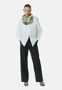 Biała koszula z guzikami, czarne spodnie o szerokich nogawkach oraz wzorzysta chusta w kolorach żółtym i szarym. Czarne sandały na paski dodają eleganckiego akcentu.