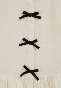 Tela color crema con tres lazos de cinta negra distribuidos de manera uniforme en vertical por el centro, encima de un dobladillo fruncido.