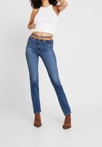 Jeans in denim blu skinny con vita alta, abbinati a una cintura marrone con fibbia rotonda. Indossati con un top corto bianco a halter e stivaletti alla caviglia marroni.