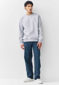 Grå sweatshirt med ribbad mudd och fåll, kombinerad med mörkblå jeans och vita sneakers, person som står mot en neutral bakgrund.