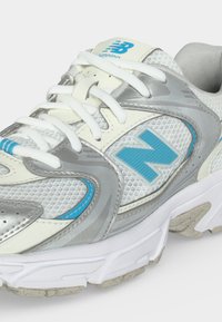 Λευκό και γκρι παπούτσι New Balance για τρέξιμο με μπλε λογότυπο, πλέγμα στο πάνω μέρος και λευκά κορδόνια, εμφανίζεται από πλάγια και πάνω γωνία.
