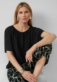 Zwart blouse met korte mouwen en geplooide halslijn, gedragen met gestreepte zwarte broek met gouden bloemenpatronen. Accenten van zilveren manchet armband.
