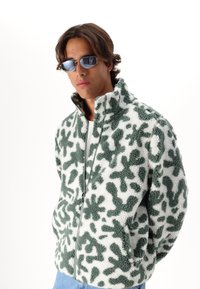 Veste en polaire verte et blanche avec un motif abstrait, col montant, fermeture éclair et poches latérales. Modèle portant des lunettes de soleil réfléchissantes.