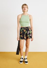 Zöld bordázott crop top fekete virágmintás rövidnadrággal párosítva. A modellen fekete sportcipő és fekete táska van. Sárga háttér.
