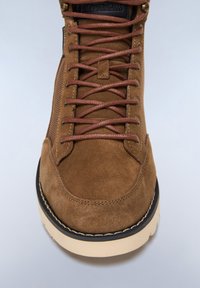 Stivale in suede marrone con lacci marroni, punta rotonda e pannello laterale testurizzato, caratterizzato da una suola color crema in contrasto e bordi neri.
