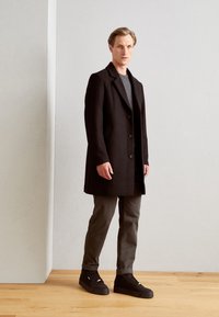 Czarny wełniany płaszcz z ostrym kołnierzem i trzema guzikami, zestawiony z szarymi spodniami i czarnymi trampkami, na tle minimalistycznego tła.