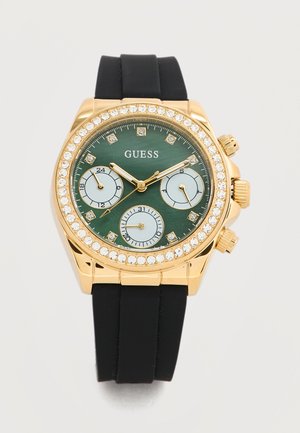 Ura Guess v zlati barvi z zelenim številčnico, tremi podštevilčnicami, kristalnimi oznakami in črnim silikonskim paščkom.