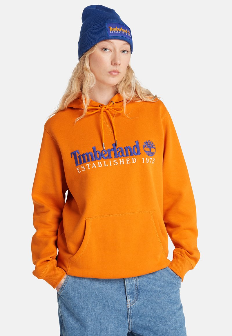 Giovane donna che indossa un vivace hoodie arancione Timberland e un berretto blu con il logo Timberland, con le mani nelle tasche dei jeans blu.