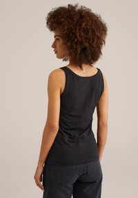 Nainen, jolla on kiharat hiukset, pukeutuneena mustaan ilman hihoja olevaan toppiin ja tummiin farkkuihin, kääntynyt hieman sivuun tasaisen taustan edessä.