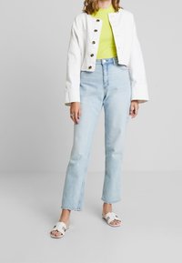 Weiße kurze Jacke mit goldenen Knöpfen über einem leuchtend grünen Rippenshirt, kombiniert mit hellblauen geraden Jeans und weißen offenen Sandalen.