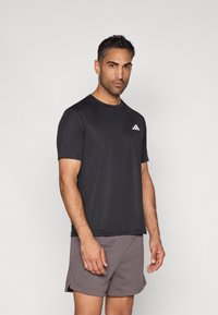 Svart trenings-T-skjorte med korte ermer, rund hals og subtil hvit Adidas-logo; kombinert med lysegrå mesh-shorts.