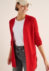 Rot gestrickte Strickjacke mit lockerer Passform, offener Vorderseite und zwei Seitentaschen. Getragen über einem weißen T-Shirt und kombiniert mit grauen Jeans.