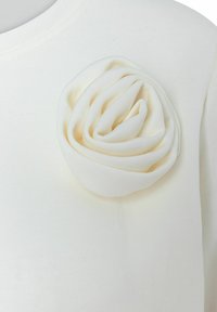 Witte gebreide top met een textured stof en een stoffen rozen detail op de linker schouder. De roos is in tinten van gebroken wit.