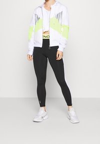 Chaqueta blanca con cremallera y acentos en verde neón y plata, un top corto blanco, leggins negros y zapatillas blancas. Tela suave, ajuste atlético.