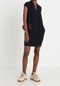 Woman wearing a sleeveless black dress, beige chunky sneakers, and a small brown shoulder bag, standing against a plain white background.