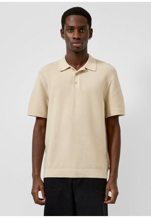 Man draagt een beige gebreid poloshirt met korte mouwen en zwarte broek, staand tegen een effen grijze achtergrond.
