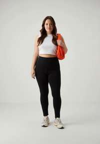 Leggings neri, canotta bianca corta e sneakers grigie. Il modello tiene una borsa arancione. Texture morbide, design semplice e colori neutri.