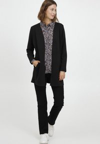 Cardigan longline nero realizzato in tessuto texturizzato, indossato su una camicia a fantasia floreale multicolore e pantaloni neri, abbinato a scarpe da ginnastica bianche.