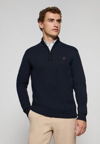 Suéter azul marino con cremallera y mangas largas, con puños y dobladillo acanalados, que presenta un pequeño logo rojo en el pecho. Llevado sobre una camisa con cuello.