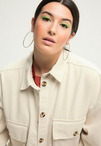 Beige, knopenjack met twee zakken, gemaakt van een gestructureerde stof. Voorzien van tortoise-shell knopen. Model heeft grote zilveren oorringen.