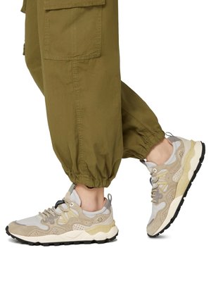 Leichte, beige Sneaker mit einem Obermaterial aus Wildleder und Mesh, gepolsterter Sohle, schwarzem Profil und dekorativen Nähten. Kombiniert mit grünen, elastischen Knöchelhosen.