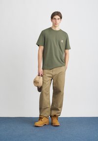 Khaki kalhoty, olivově zelené tričko s kapsou na prsou, béžová kšiltovka a žluté boty. Jednoduchý design s praktickými, neformálními prvky.