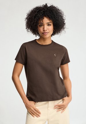 Jonge vrouw met krullend haar, gekleed in een donkerbruin shirt met korte mouwen en licht beige broek, staande met de handen nabij de zakken tegen een effen achtergrond.