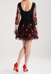 Černé šaty s průhlednými tříčtvrtečními rukávy, ozdobené červenou květinovou výšivkou. Zadní strana má hluboký výstřih a zapínání na zip.