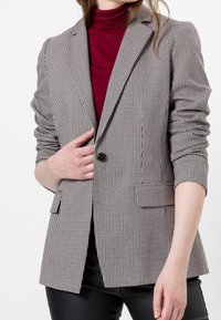 Blazer à motif pied-de-poule en noir et blanc, avec un seul bouton, des revers crantés et deux poches avant, porté sur un haut bordeaux.