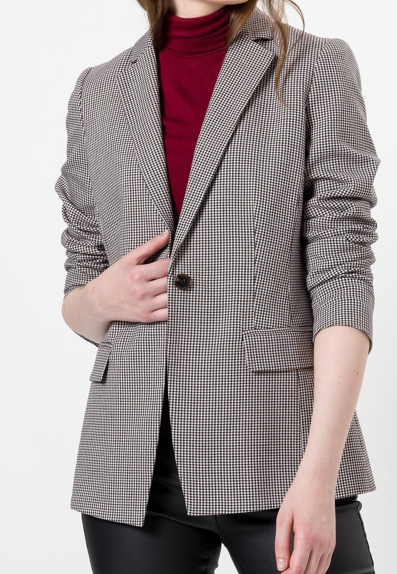 Blazer à motif pied-de-poule en noir et blanc, avec un seul bouton, des revers crantés et deux poches avant, porté sur un haut bordeaux.