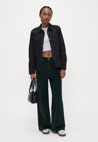 Zwarte spijkerjack met gestikte schouderaccenten, grijze cropped top, donkergroene corduroy wijde broek en zwarte handtas. Witte sneakers.