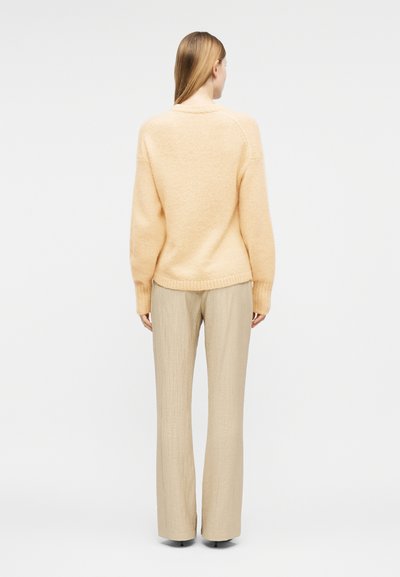 Femme debout de dos, portant un pull en tricot pêche et un pantalon beige à jambes larges, sur un fond blanc uni.