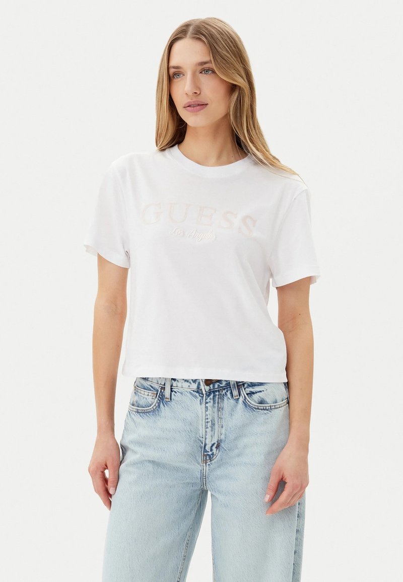 T-shirt bianca cropped con logo "GUESS Los Angeles" in morbido ricamo rosa, caratterizzata da un classico collo rotondo e maniche corte.