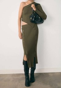 Frau in einem olivgrünen, gerippten Strick-Zweiteiler mit Knopfdetails, kniehohen schwarzen Stiefeln und einer schwarzen Handtasche an einer Schulter.