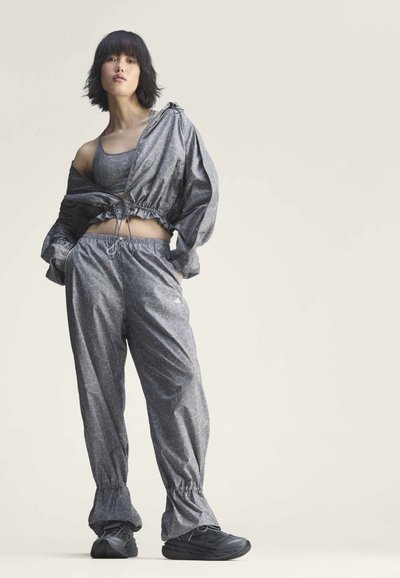 Jeune femme portant un soutien-gorge de sport gris à motif floral assorti, une veste, un pantalon et des baskets noires, debout, les mains dans les poches.