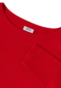 Camicetta rossa in cotone a maniche lunghe con scollatura rotonda. Presenta un'etichetta che recita "CEcil" vicino al collo e una superficie liscia.