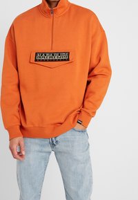 Orange pullover with a high collar, featuring a zip and a stitched black patch with "NAPAPURI geographic." Cotton blend, relaxed fit.