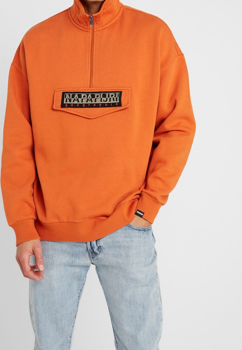 Orange pullover with a high collar, featuring a zip and a stitched black patch with "NAPAPURI geographic." Cotton blend, relaxed fit.