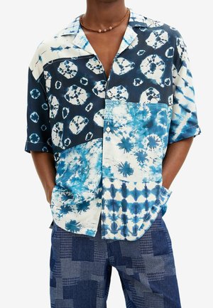 Mężczyzna ubrany w koszulę na guziki z krótkim rękawem w niebiesko-białe wzory tie-dye oraz w niebieskie spodnie z łatającym wzorem, z rękami w kieszeniach.