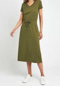 Robe verte olive longueur genoux en tissu doux avec manches courtes, décolleté en V, taille réglable avec cordon, portée avec des baskets blanches à plateforme.