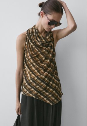 Kobieta w czarnych okularach przeciwsłonecznych i brązowej, kraciastej, bezrękawnikowej bluzce, trzymająca czarną torebkę, na tle jednolitego, szarego tła.