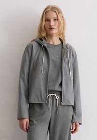 Szara bluza z kapturem zapinana na zamek, z troczkami i przednią kieszenią, zestawiona z szarą koszulką z okrągłym dekoltem, prezentowana na modelu.