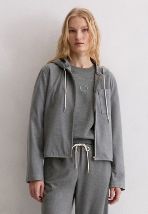 Felpa grigia con zip, cappuccio, coulisse e tasca frontale, abbinata a una maglia grigia girocollo, indossata da un modello.