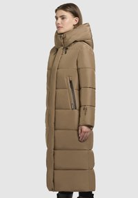 Dlhý béžový puffový kabát s polstrovaným dizajnom, nastaviteľnou kapucňou a dvojitými zipsovými vreckami, vystupujúci elegantnou, hladkou textúrou a priamym strihom.
