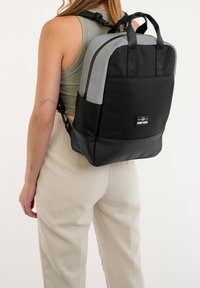 Backpack in black and gray, made of durable material. Features two top handles, adjustable straps, and a front label. Smooth texture.