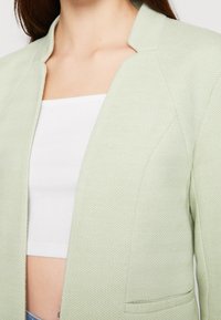 Blazer verde claro con un sutil patrón espiga, con cuello chal y frontal abierto. El material es texturizado y de diseño ajustado.