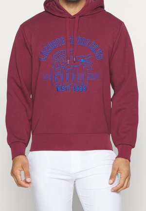 Bordeauxroter Kapuzenpullover mit blauem Schriftzug „Lacoste Sport Club Golf Est. 1933“ und Krokodil-Logo, getragen zu weißen Hosen.