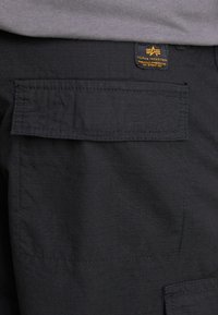 Detail z blízka na čiernu kapsu na cargo nohaviciach s chlopňou a označením značky Alpha Industries nad kapsou v oblasti pásu.