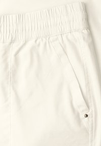Détail d’un pantalon en tissu blanc montrant une taille élastique et une poche latérale avec un petit bouton métallique.