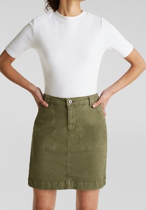 Vrouw draagt een effen wit T-shirt met korte mouwen en een olijfgroene rok met knopen en voorzakken, handen op de heupen, neutrale achtergrond.